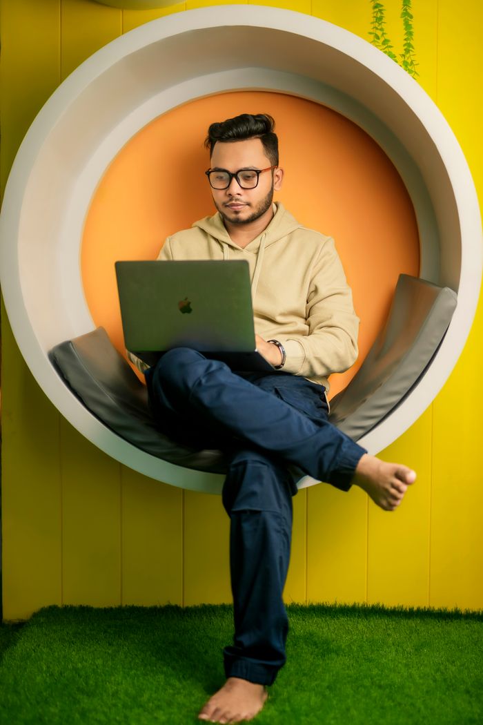 Young adult man using a laptop while seated in a modern circular chair, set against a vibrant background.